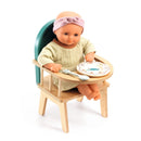 The Djeco Baby Chair features a feeding doll dressed in a pink headband and green outfit sitting in the wooden chair, crafted from FSC certified wood with a green backrest. In front of the doll is a small plate and spoon, perfect for mealtime magic.