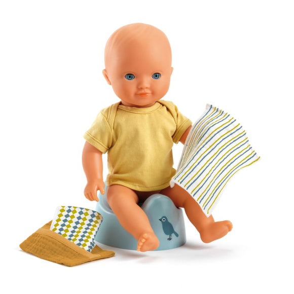 A Djeco baby doll sits on the blue Djeco Doll Potty adorned with a bird design, wearing a yellow shirt and holding a striped cloth. Nearby is an FSC-certified wooden doll chair with a matching hat.