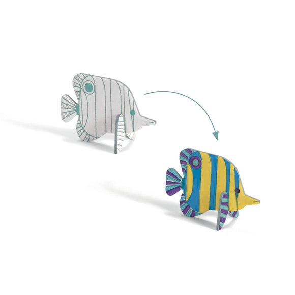 Djeco's "Colour, Assemble & Play Sea-Life" kit includes two FSC certified cardboard fish models: one uncolored with outlined stripes and the other vibrantly decorated in yellow, blue, and purple. An arrow showcases the magic of transforming sea-life through coloring and assembling.