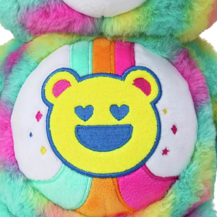 Close-up of the 35cm Care Bears Eco-friendly Good Vibes Bear by Basic Fun, showcasing vibrant rainbow colors with a yellow bear face, heart-shaped eyes, and a wide smile embroidered on its belly. The background has fluffy textures in light blue, pink, and green.