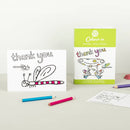 A handmade thank you card with a colorful dragonfly design from Eggnog's "Card Book - Colour-in Thank You Cards" is showcased with its packaging. Nearby are three colored pencils, an envelope, and a handwritten note.