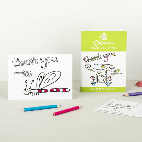 A handmade thank you card with a colorful dragonfly design from Eggnog's "Card Book - Colour-in Thank You Cards" is showcased with its packaging. Nearby are three colored pencils, an envelope, and a handwritten note.