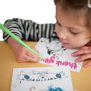 A brown-haired child wearing a striped shirt uses a green marker from the Eggnog Card Book - Colour-in Thank You Cards to create a thank you card featuring a pink "Thank you" and a monster drawing. Another card with blue and brown scribbles lies on the table.
