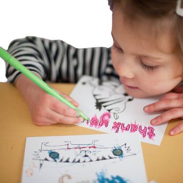 A brown-haired child wearing a striped shirt uses a green marker from the Eggnog Card Book - Colour-in Thank You Cards to create a thank you card featuring a pink "Thank you" and a monster drawing. Another card with blue and brown scribbles lies on the table.