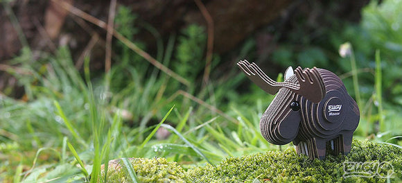 The EUGY Moose by EUGY is a small, artistic sculpture made from sustainable materials, set on moss-covered terrain amidst lush green grass. Its intricate design features delicately cut pieces with detailed antlers, and the blurred background enhances the focus on this eco-friendly masterpiece.