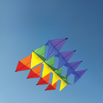The SPIRIT OF AIR Aurora Box Kite features vibrant multi-tier triangular sails in red, orange, yellow, green, and purple. Its sturdy glass fiber frame ensures effortless flying against a clear blue sky for enthusiasts of all ages.