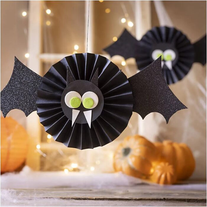 Two Halloween bats from the Creativ Company Mini Craft Kit Paper Decorations hang in front of a fall-themed backdrop with pumpkins and warm string lights, featuring black paper bodies with green and white eyes and white fangs.