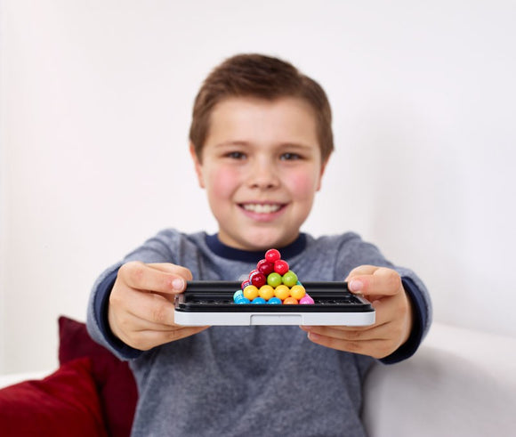 A cheerful child sits on a white couch with a red cushion, showcasing the IQ Puzzler Pro by Smart Games—a colorful pyramid of spheres—towards the camera, enjoying some brain-teasing fun.