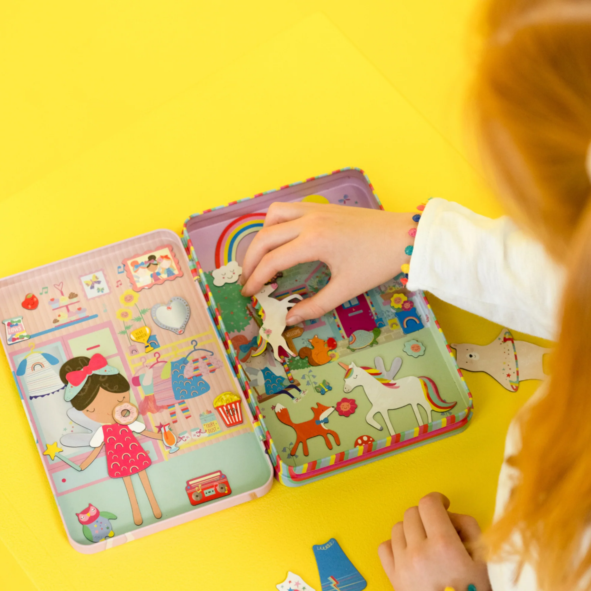 A child joyfully engages with the Rainbow Fairy Magnetic Playtime set from Floss & Rock on a bright yellow surface. The vibrant, eco-friendly pieces, crafted from recycled materials, feature unicorns and other delightful elements. While assembling pop-out magnets on the colorful magnetic tin case, the child wears a bracelet.