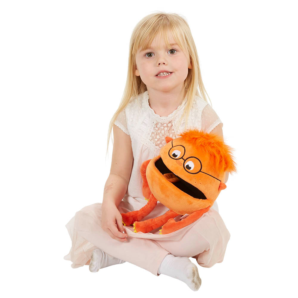 A young blonde girl sits on the floor in a white and light pink dress, holding an Orange Baby Monster Puppet from THE PUPPET COMPANY, featuring big eyes, glasses, and an open mouth ready for chat with its squeaky voice.