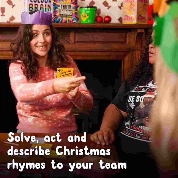 A woman wearing a purple paper crown and a festive sweater holds a card while smiling, seated at a table with others. Text at the bottom reads, "Solve, act, and describe Christmas rhymes to your team." Festive decorations are in the background, setting the perfect scene for playing Santa Banter by The Toy Shop—an entertaining Christmas game.