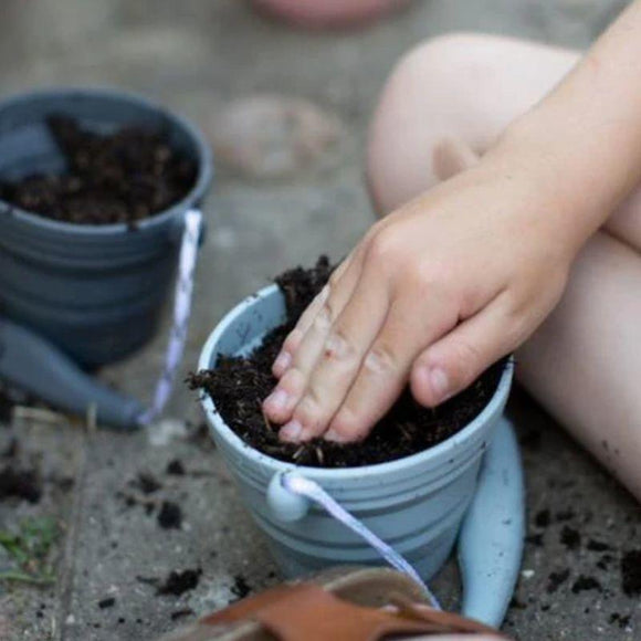 A child in shorts uses a SCRUNCH Mini Bucket & Spade to press soil into a small gray pot. Another collapsible pot is visible in the background, while gardening tools and soil surround the young gardener.