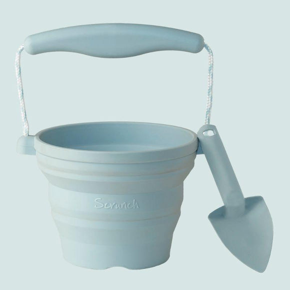 The Scrunch Mini Bucket & Spade by SCRUNCH in Duck Egg features a silicone bucket with a flexible handle and matching small shovel, ideal for little gardeners. "Scrunch" is embossed on the side, set against a soft blue backdrop.
