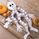 A paper skeleton from the Creativ Company Mini Craft Kit Paper Decorations set sits on a wooden surface, assembled with white paper chains. Beside it are mini pumpkins, rolled construction paper, and a glue stick. Spider and web patterns embellish its hands and feet, making it an ideal Halloween decoration.