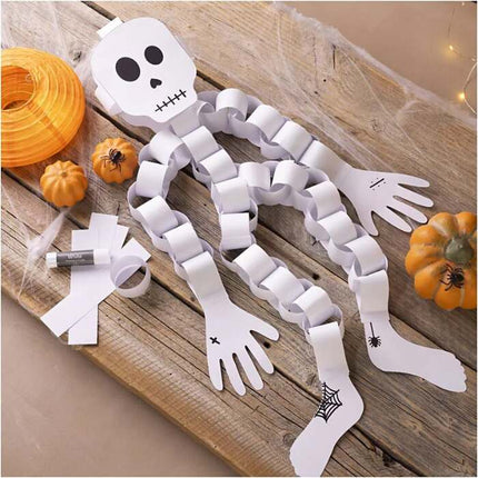 A paper skeleton from the Creativ Company Mini Craft Kit Paper Decorations set sits on a wooden surface, assembled with white paper chains. Beside it are mini pumpkins, rolled construction paper, and a glue stick. Spider and web patterns embellish its hands and feet, making it an ideal Halloween decoration.