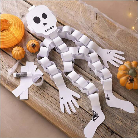 A paper skeleton from the Creativ Company Mini Craft Kit Paper Decorations set sits on a wooden surface, assembled with white paper chains. Beside it are mini pumpkins, rolled construction paper, and a glue stick. Spider and web patterns embellish its hands and feet, making it an ideal Halloween decoration.