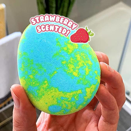 A hand holds a Baff Egg Bombz Green & Blue Turtle by Zimpli Kids, showcasing blue and yellow swirls. A digital overlay reads "STRAWBERRY SCENTED!" with an illustrated strawberry. Experience a fizzing bath adventure that's vegan-friendly, with a blurred bathroom in the background.