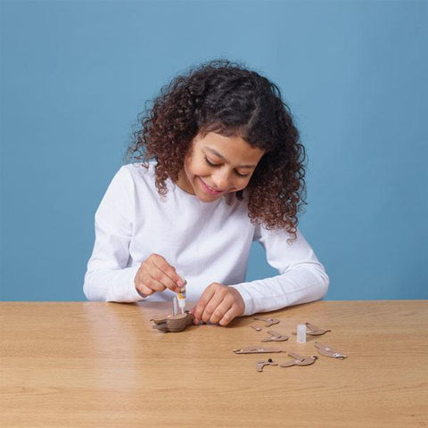 Girl with dark hair and long sleeved white t-shirt sitting down at a table and glueing pieces of the eugy sloth figure.