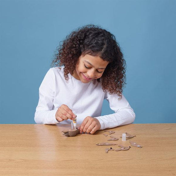 Girl with dark hair and long sleeved white t-shirt sitting down at a table and glueing pieces of the eugy sloth figure.