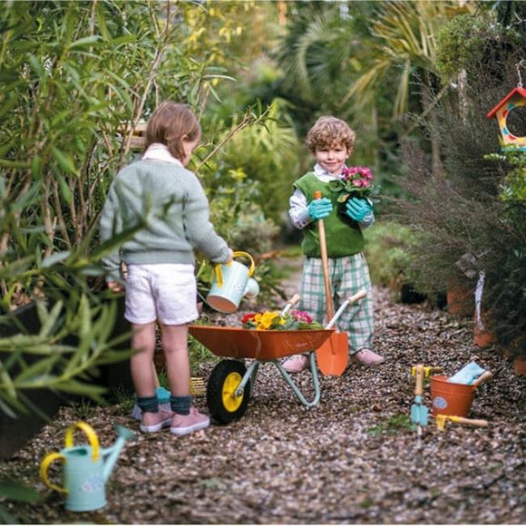 Two children playing on a garden path with the orange wheelbarrow and other gardeing toys.