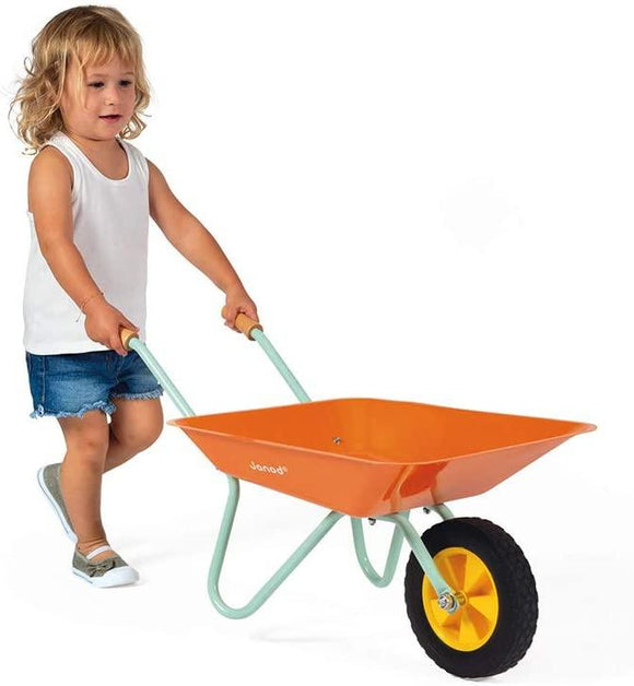 Young girl with blonde hair, denim skirt and white top pushing the orange wheelbarrow.