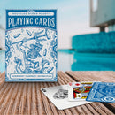 Displayed on a wooden surface by a pool, the Ocean Plastic Playing Cards from Big Potato Games showcase an eco-friendly design made from recycled ocean plastic. The waterproof box features a blue color scheme with an illustration of a bearded figure holding a trident, surrounded by ocean-themed elements.