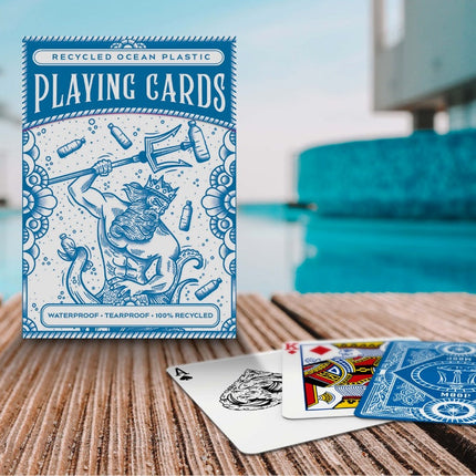 Displayed on a wooden surface by a pool, the Ocean Plastic Playing Cards from Big Potato Games showcase an eco-friendly design made from recycled ocean plastic. The waterproof box features a blue color scheme with an illustration of a bearded figure holding a trident, surrounded by ocean-themed elements.