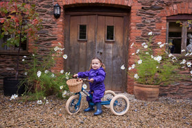 Child wearing a purple rainsuit on a green Trybike in front of a red brick house.