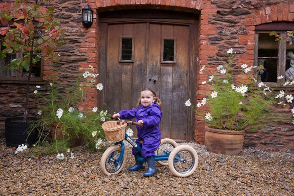 Child wearing a purple rainsuit on a green Trybike in front of a red brick house.